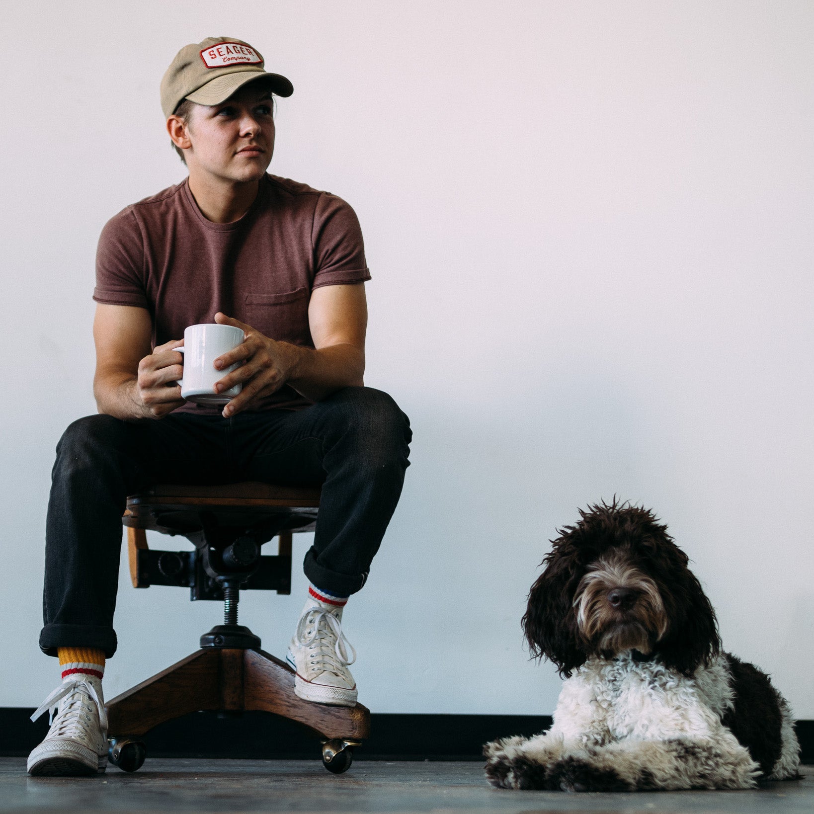 Man in "Seager Company" cap enjoys coffee with his dog.
