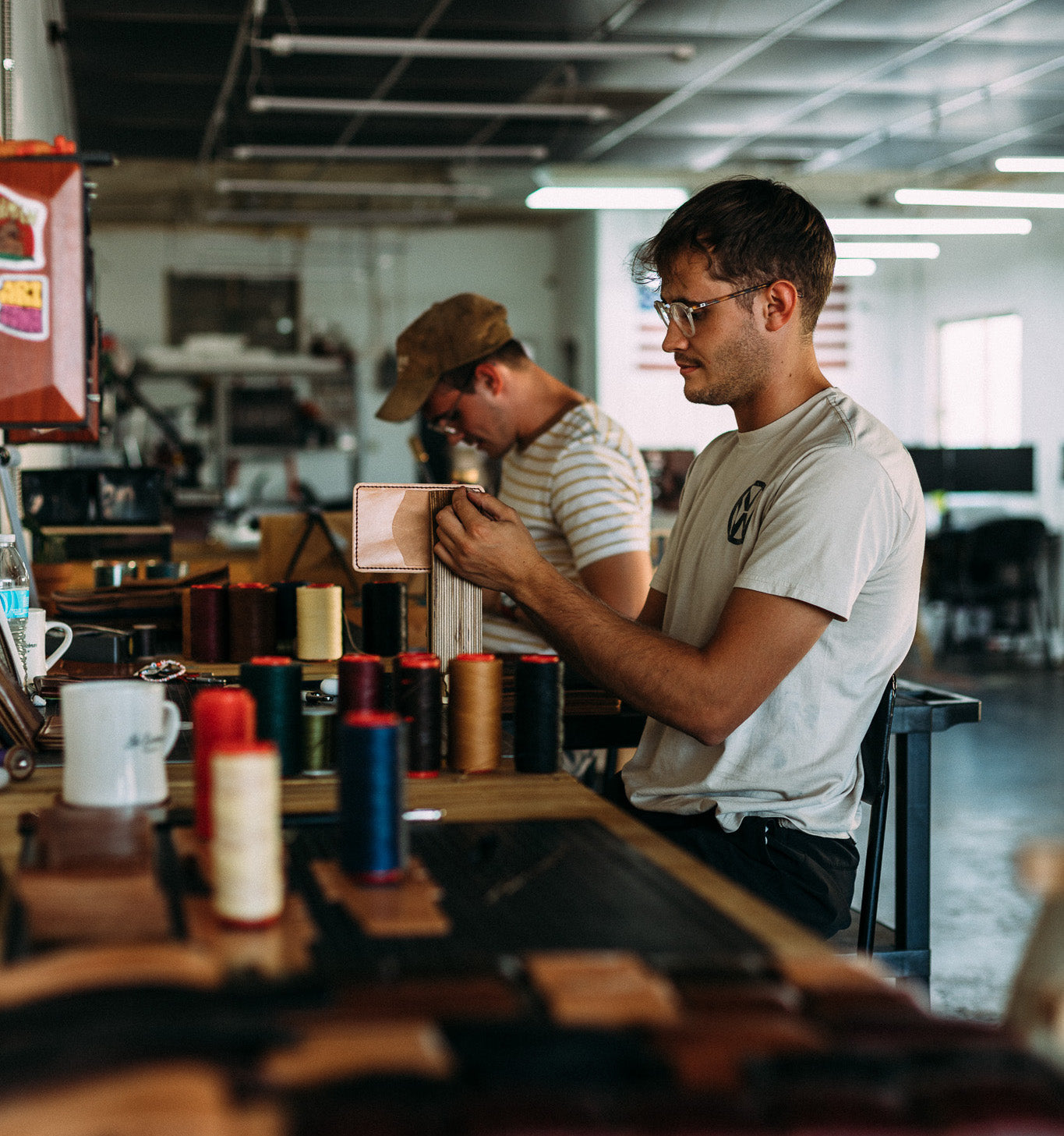 Leatherworkers handcrafting a wallet with spools of thread in a workshop.