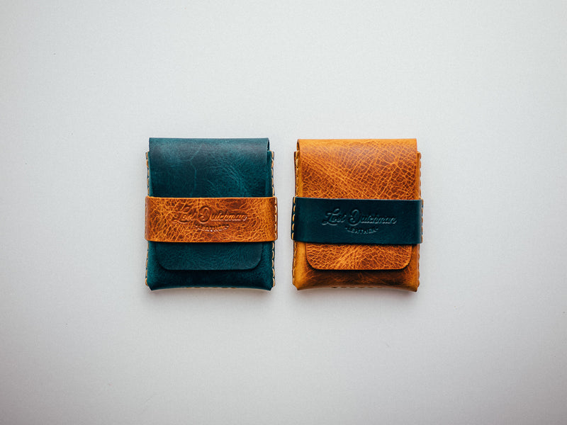 Two dark teal and brown leather card holders on a white surface